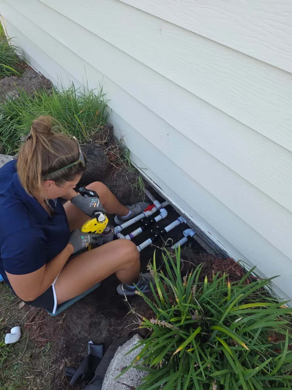 IrriGators LLC technician cutting PVC pipe to rebuild a sprinkler valve manifold beside a home