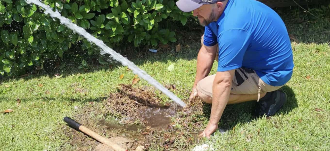 IrriGators LLC technician kneeling beside a broken sprinkler line spraying water across a lawn