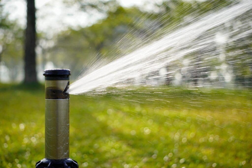 Sprinkler spraying water onto a green lawn, blurred background of trees.